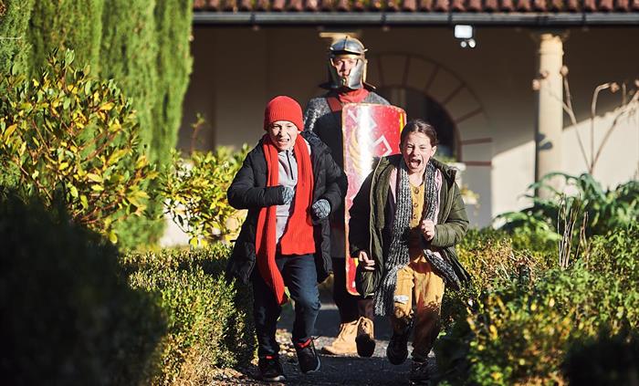 two children run along a gravel path, flanked by low hedges. in the background, there's a man dressed in Roman armour carrying a red shield.